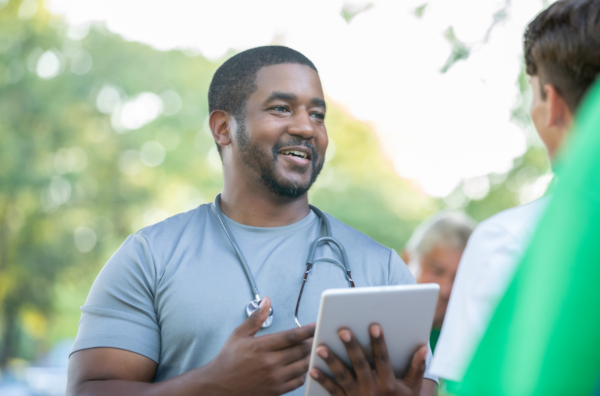 A Black man with a tablet and stethoscope speaking with another person to collect data for a program evaluation
