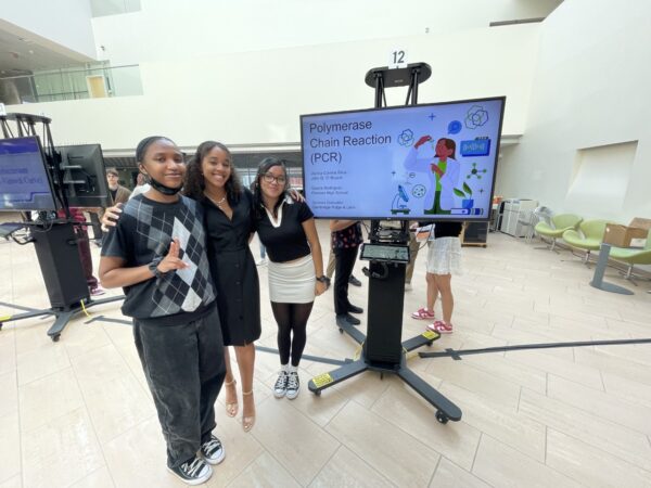 Three young people in a school gym standing next to a screen displaying a presentation entitled "Polymerase Chain Reaction (PCR)"